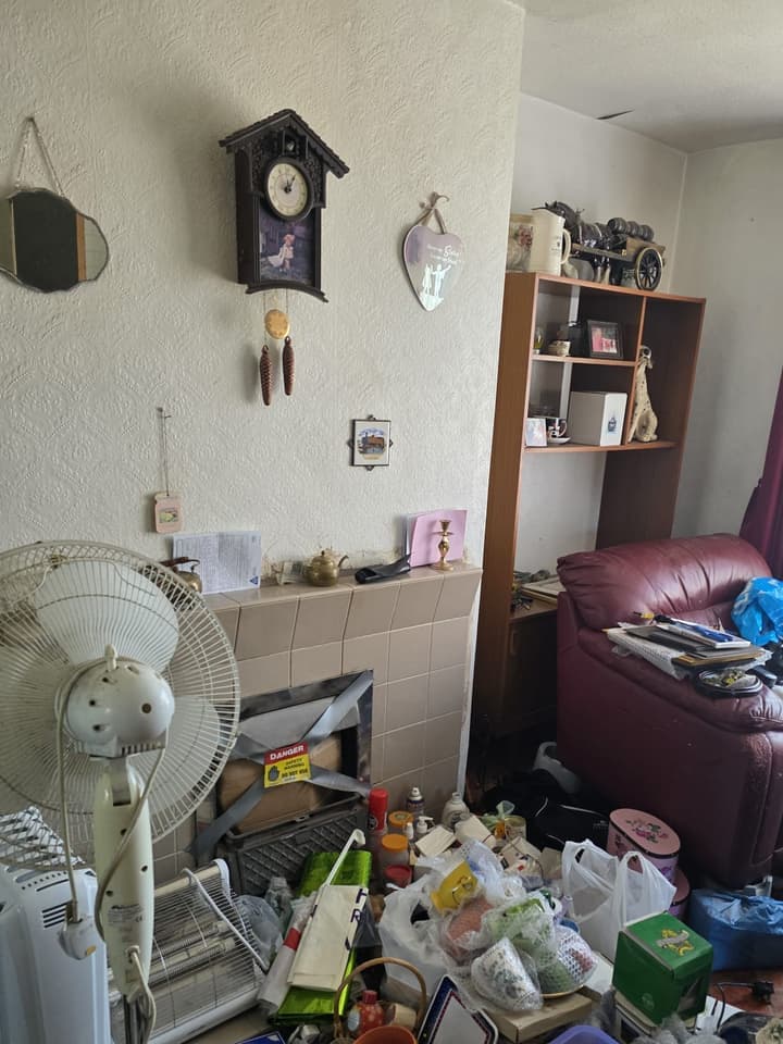 Living room before the clearance: a fan, a clock on the wall, bags and belongings across the floor